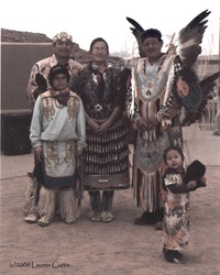 Photograph in faded colors of a Native American family living in the Grand canyon area of Arizona. They are dressed in ceremonail, decorative clothing with beads and feathers. Several generations are posing here.