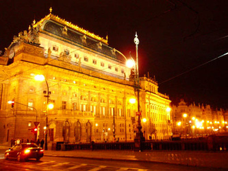 Photo of an historic building, lit up at night, in down town Prague, Czech Republic.  Bright yellows and oranges from the lights. Ornate architecture.